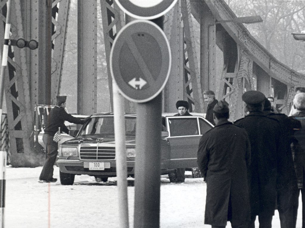 Im Vordergrund sind einige, teilweise uniformierte Männer mit dem Rücken zur Kamera und ein Verkehrsschild zu sehen. Zentral steht eine Limousine auf der Glienicker Brücke. An der geöffneten linken Hintertür stehen Anatoli Schtscharanski und ein weiterer Mann, ein anderer öffnet die rechte Vordertür.