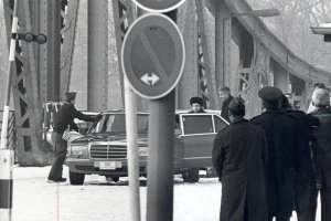 Im Vordergrund sind einige, teilweise uniformierte Männer mit dem Rücken zur Kamera und ein Verkehrsschild zu sehen. Zentral steht eine Limousine auf der Glienicker Brücke. An der geöffneten linken Hintertür stehen Anatoli Schtscharanski und ein weiterer Mann, ein anderer öffnet die rechte Vordertür.