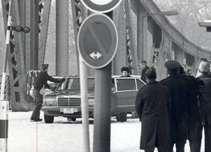Im Vordergrund sind einige, teilweise uniformierte Männer mit dem Rücken zur Kamera und ein Verkehrsschild zu sehen. Zentral steht eine Limousine auf der Glienicker Brücke. An der geöffneten linken Hintertür stehen Anatoli Schtscharanski und ein weiterer Mann, ein anderer öffnet die rechte Vordertür.
