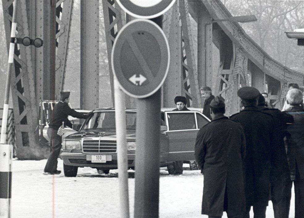 Im Vordergrund sind einige, teilweise uniformierte Männer mit dem Rücken zur Kamera und ein Verkehrsschild zu sehen. Zentral steht eine Limousine auf der Glienicker Brücke. An der geöffneten linken Hintertür stehen Anatoli Schtscharanski und ein weiterer Mann, ein anderer öffnet die rechte Vordertür.