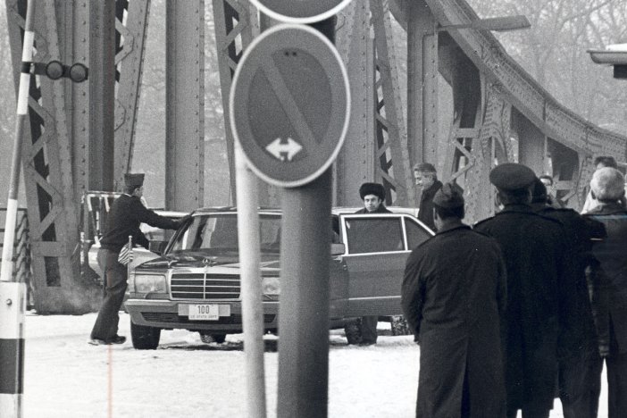 Im Vordergrund sind einige, teilweise uniformierte Männer mit dem Rücken zur Kamera und ein Verkehrsschild zu sehen. Zentral steht eine Limousine auf der Glienicker Brücke. An der geöffneten linken Hintertür stehen Anatoli Schtscharanski und ein weiterer Mann, ein anderer öffnet die rechte Vordertür.