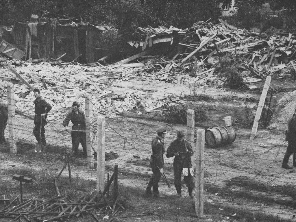 Im Vordergrund stehen mehrere uniformierte Männer zwischen Stacheldrahtverhauen. Im Hintergrund sind Haufen von Holzlatten und halbzerstörte Hütten zu sehen.