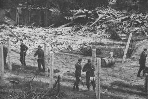 Im Vordergrund stehen mehrere uniformierte Männer zwischen Stacheldrahtverhauen. Im Hintergrund sind Haufen von Holzlatten und halbzerstörte Hütten zu sehen.