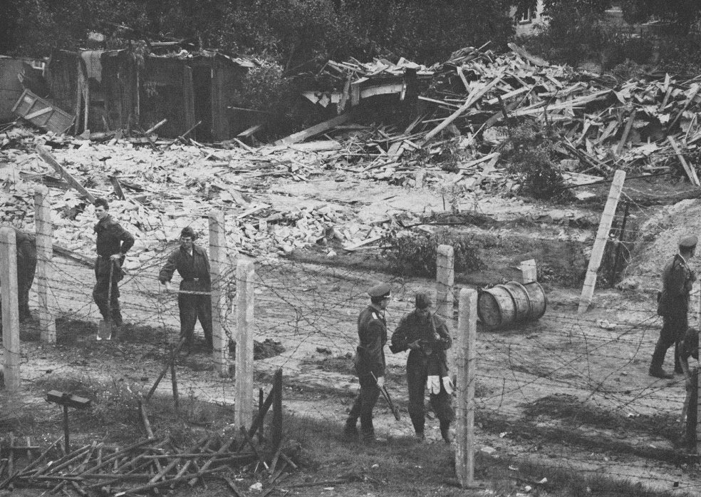 Im Vordergrund stehen mehrere uniformierte M&auml;nner zwischen Stacheldrahtverhauen. Im Hintergrund sind Haufen von Holzlatten und halbzerst&ouml;rte H&uuml;tten zu sehen.