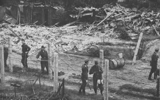 Im Vordergrund stehen mehrere uniformierte Männer zwischen Stacheldrahtverhauen. Im Hintergrund sind Haufen von Holzlatten und halbzerstörte Hütten zu sehen.