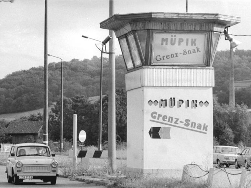 Links fährt ein Trabbi auf die Kamera zu. Rechts steht ein Wachturm, in dessen Fenster ein Plakat verkündet, Müpik, Grenz Snack. Der Turm ist mit derselben Aufschrift bemalt.