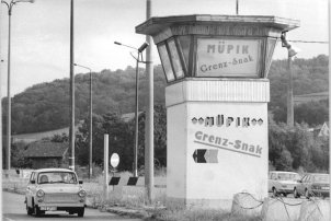 Links fährt ein Trabbi auf die Kamera zu. Rechts steht ein Wachturm, in dessen Fenster ein Plakat verkündet, Müpik, Grenz Snack. Der Turm ist mit derselben Aufschrift bemalt.