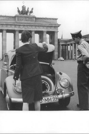 Eine Frau steht mit dem Rücken zur Kamera vor einem VW-Käfer und öffnet den Kofferraum. Rechts daneben steht ein Uniformierter und schaut hinein. Im Hintergrund ist das Brandenburger Tor zu sehen.