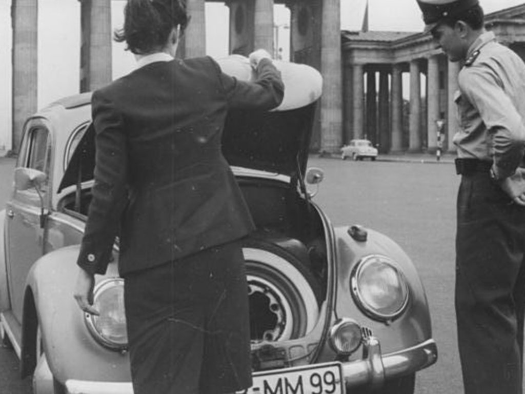 Eine Frau steht mit dem Rücken zur Kamera vor einem VW-Käfer und öffnet den Kofferraum. Rechts daneben steht ein Uniformierter und schaut hinein. Im Hintergrund ist das Brandenburger Tor zu sehen.