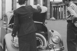 Eine Frau steht mit dem Rücken zur Kamera vor einem VW-Käfer und öffnet den Kofferraum. Rechts daneben steht ein Uniformierter und schaut hinein. Im Hintergrund ist das Brandenburger Tor zu sehen.