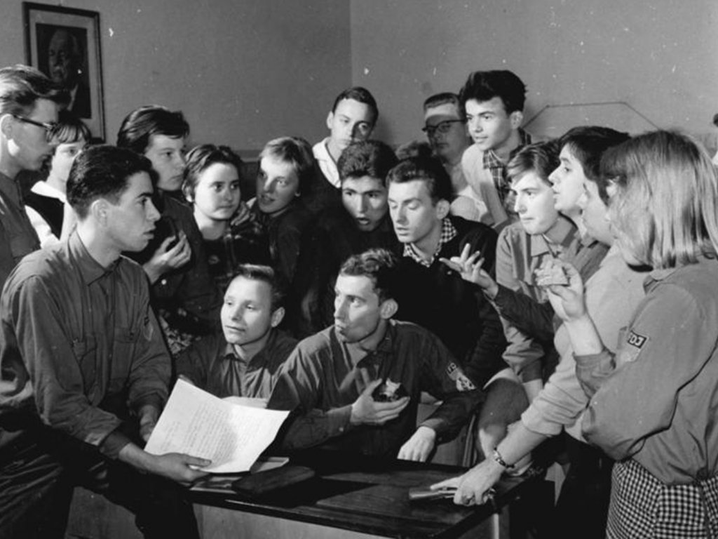 FDJ-Gruppe bei der „Diskussion“ über einen Aufruf gegen das Westfernsehen und das Hören von „NATO-Rundfunksendern“; Aufnahme 26. September 1961 Eine Gruppe junger Menschen steht um einen Tisch, redet und gestikuliert. Einige tragen FDJ-Hemden. Links im Vordergrund sitzt ein junger Mann mit einem Bein auf dem Tisch und hält ein beschriebenes Papier in der Hand. Im Hintergrund hängt ein Porträt von Wilhelm Pieck an der Wand.