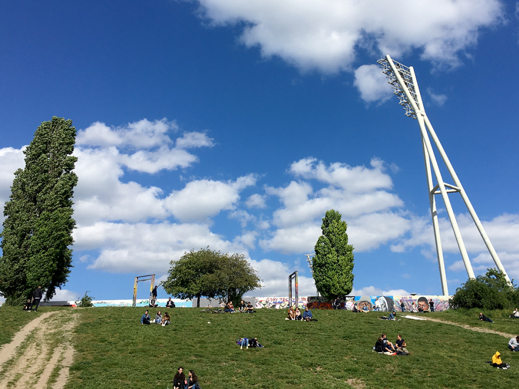 Auf einer grünen Anhöhe sitzen Menschen in kleinen Gruppen zusammen. Oben auf dem Hügel, nur klein zu erkennen, steht ein bemaltes Stück der Hinterlandmauer. Einige Bäume und die Flutlichtanlage des Friedrich Ludwig Jahn Sportparks ragen hoch in den blauen Himmel.
