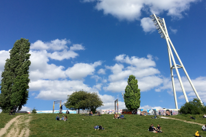 Auf einer grünen Anhöhe sitzen Menschen in kleinen Gruppen zusammen. Oben auf dem Hügel, nur klein zu erkennen, steht ein bemaltes Stück der Hinterlandmauer. Einige Bäume und die Flutlichtanlage des Friedrich Ludwig Jahn Sportparks ragen hoch in den blauen Himmel.