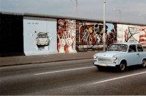 Abschnitt der East Side Gallery mit den Bildern Von Hunter und Russel im Hintergrund. Vorne im Bild ist das Gemälde eines Trabis, der durch die Mauer bricht. Über dem Trabi steht: Test The Best. Auf der Straße im Vordergrund fährt ein Trabi.