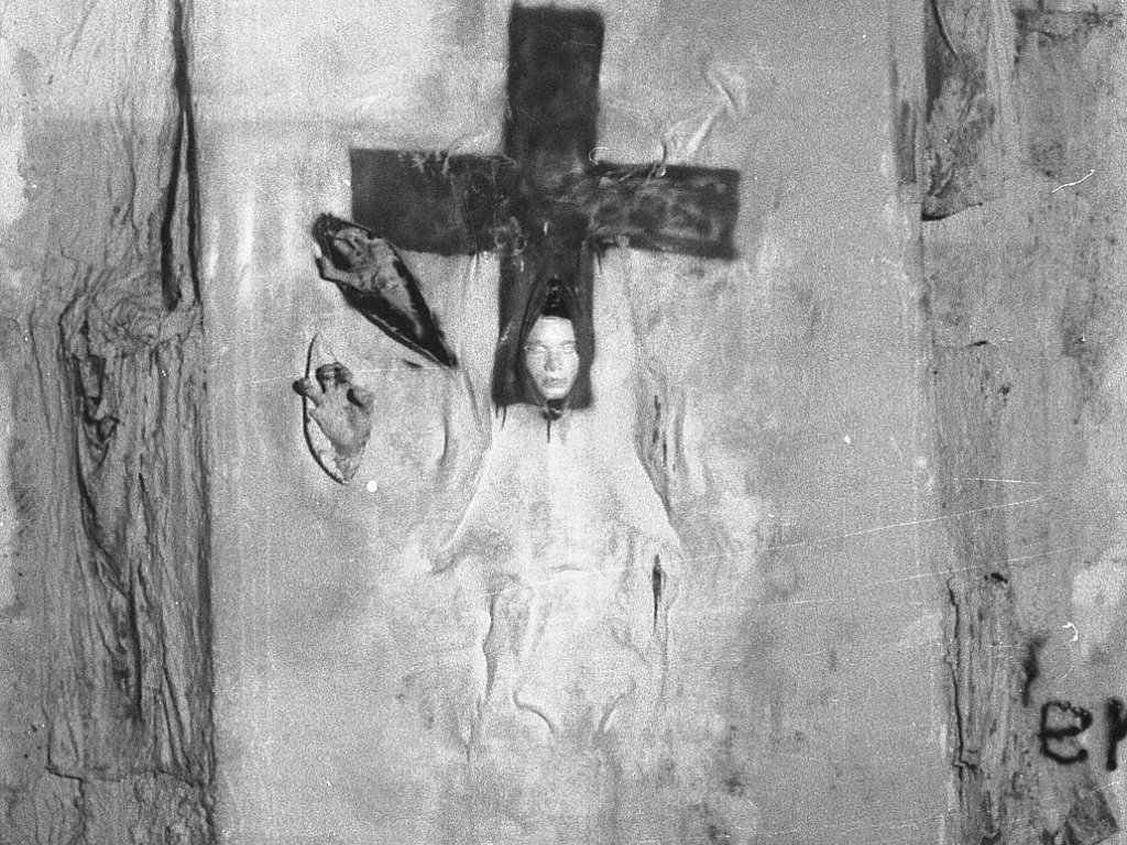 Mauerabschnitt mit der Installation. Ein Tuch ist auf der Mauer angebracht, darauf ist im oberen Bereich ein Kreuz gemalt. Aus dem unteren Teil des Kreuzes ragt eine Gipsmaske. Links neben dem Kreuz sind zwei weitere Schlitze im Stoff aus denen je eine Hand ragt.