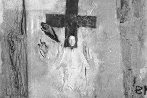 Mauerabschnitt mit der Installation. Ein Tuch ist auf der Mauer angebracht, darauf ist im oberen Bereich ein Kreuz gemalt. Aus dem unteren Teil des Kreuzes ragt eine Gipsmaske. Links neben dem Kreuz sind zwei weitere Schlitze im Stoff aus denen je eine Hand ragt.