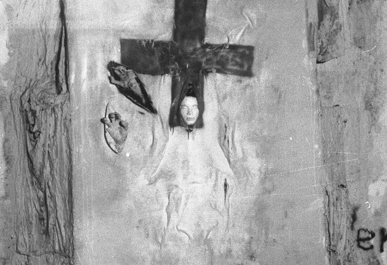 Mauerabschnitt mit der Installation. Ein Tuch ist auf der Mauer angebracht, darauf ist im oberen Bereich ein Kreuz gemalt. Aus dem unteren Teil des Kreuzes ragt eine Gipsmaske. Links neben dem Kreuz sind zwei weitere Schlitze im Stoff aus denen je eine Hand ragt.