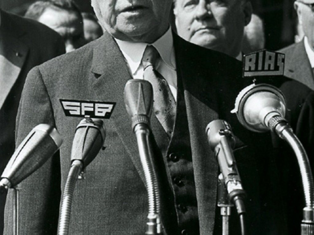 Konrad Adenauer steht mit ernstem Blick vor Mikrofonen und spricht.