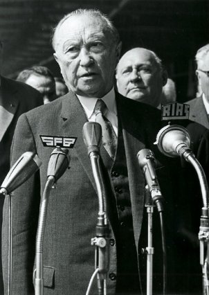 Konrad Adenauer steht mit ernstem Blick vor Mikrofonen und spricht.