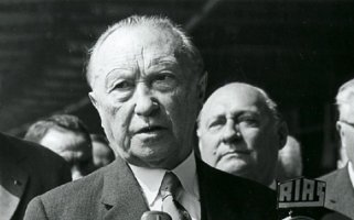 Konrad Adenauer steht mit ernstem Blick vor Mikrofonen und spricht.