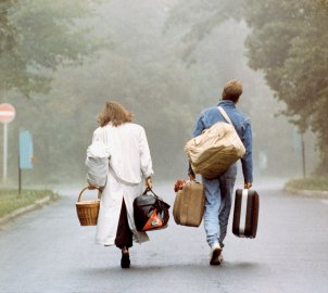 Eine Frau und ein Mann sind von hinten zu sehen, wie sie mit viel Gepäck im Nebel auf eine Straßengabelung zugehen.