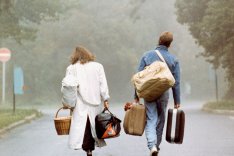 Eine Frau und ein Mann sind von hinten zu sehen, wie sie mit viel Gepäck im Nebel auf eine Straßengabelung zugehen.