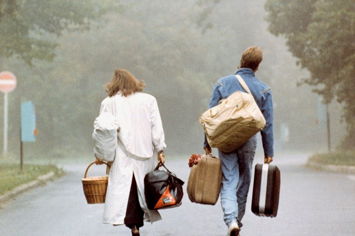 Eine Frau und ein Mann sind von hinten zu sehen, wie sie mit viel Gepäck im Nebel auf eine Straßengabelung zugehen.