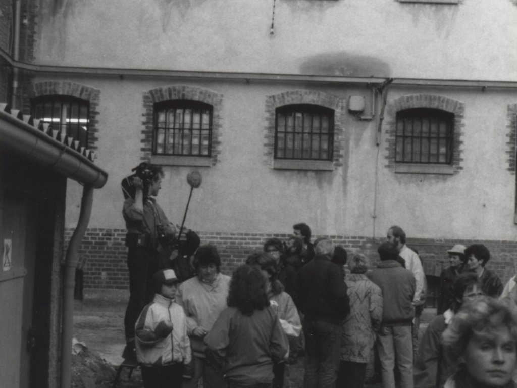 Eine Gruppe Menschen steht vor einer Hauswand mit vergitterten Fenstern. Ein Mann auf einer Trittleiter hält eine Kamera und ein Mikrofon.