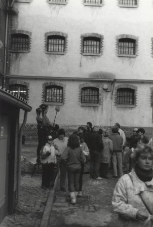 Eine Gruppe Menschen steht vor einer Hauswand mit vergitterten Fenstern. Ein Mann auf einer Trittleiter hält eine Kamera und ein Mikrofon.