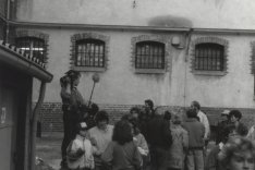 Eine Gruppe Menschen steht vor einer Hauswand mit vergitterten Fenstern. Ein Mann auf einer Trittleiter hält eine Kamera und ein Mikrofon.