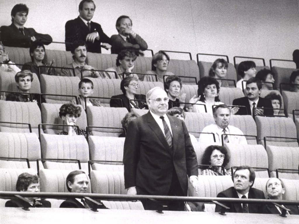 Die Sitze der Gästetribüne der Volkskammer sind zum Großteil belegt. In der vordersten Reihe steht Helmut Kohl, einige der um ihn Sitzenden schauen zu ihm auf.