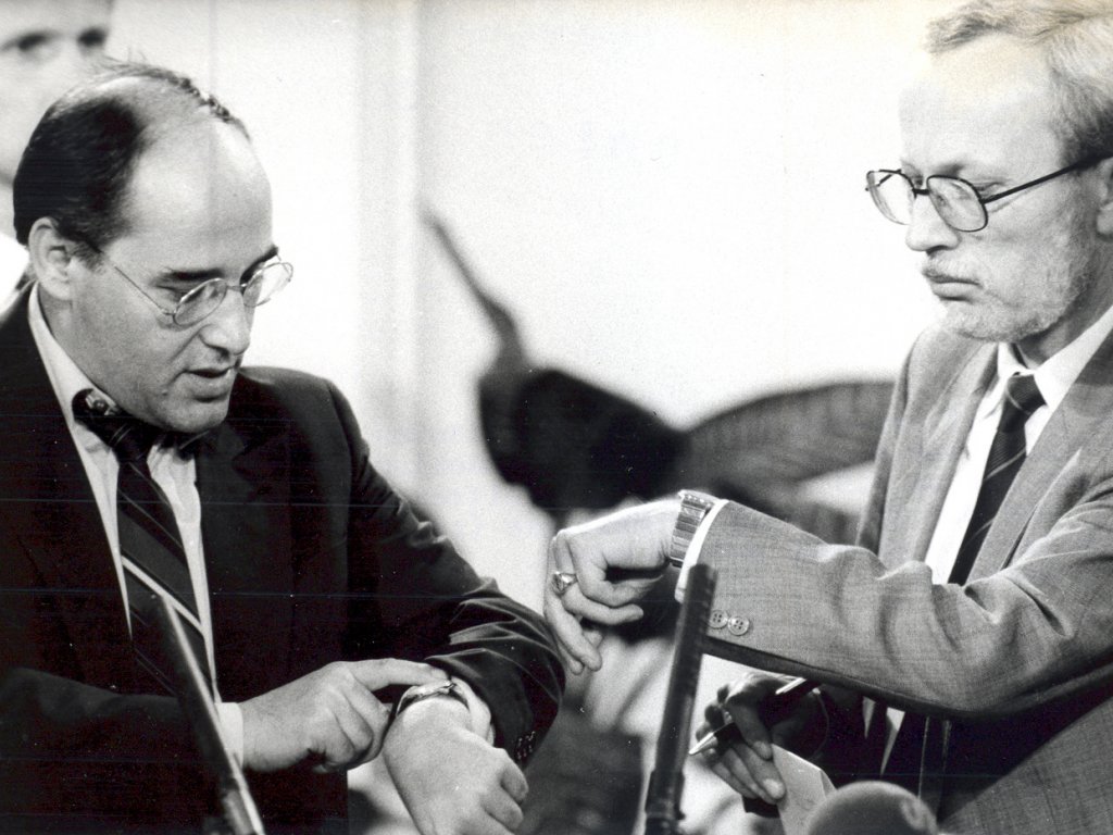 Lothar de Maizière und Gregor Gysi kurz vor Beginn einer Fernsehwahlsendung am Abend der Kommunalwahlen in der DDR, 6. Mai 1990. Gregor Gysi links und Lothar de Maizière rechts sind einander zugewandt und schauen beide auf ihre Armbanduhren. Im Vordergrund sind unscharf auf die beiden gerichtete Mikrofone zu erkennen.