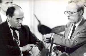 Gregor Gysi links und Lothar de Maizière rechts sind einander zugewandt und schauen beide auf ihre Armbanduhren. Im Vordergrund sind unscharf auf die beiden gerichtete Mikrofone zu erkennen.