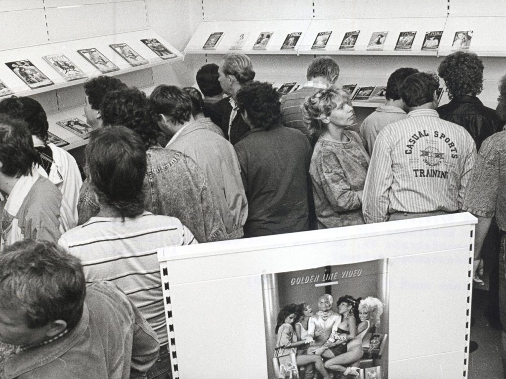 Bei der Eröffnung des ersten Sex-Shops in Cottbus ist der Andrang groß, Juli 1990. Eine Menschenmenge steht dicht gedrängt in einem Raum mit Zeitschriftenregalen an den Wänden. Im Vordergrund steht ein Aufsteller mit einem Plakat, das die Aufschrift Golden Live Video trägt. Das Plakat zeigt Bild von einem Mann, der von fünf leicht bekleideten Frauen umringt wird, die sich an ihn schmiegen.