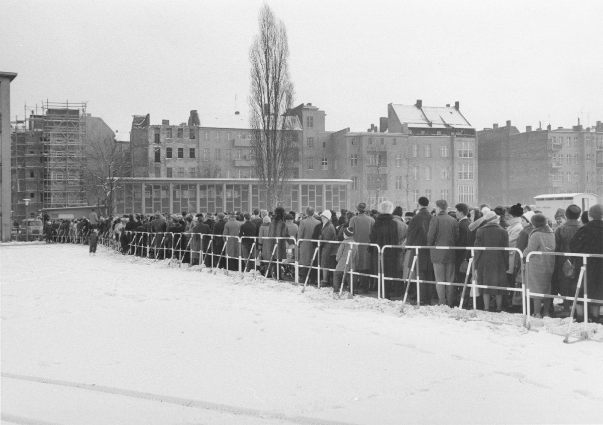 Eine sehr lange Menschenschlange steht entlang eines Absperrgitters im Schnee vor einem Gebäude, das klein im Hintergrund zu erkennen ist.