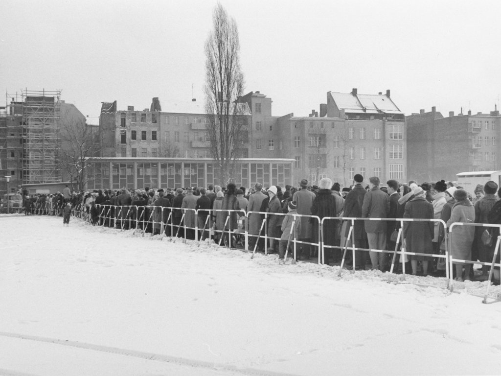 Warteschlangen für Passierscheine vor einer West-Berliner Schule, Dezember 1963 Eine sehr lange Menschenschlange steht entlang eines Absperrgitters im Schnee vor einem Gebäude, das klein im Hintergrund zu erkennen ist.
