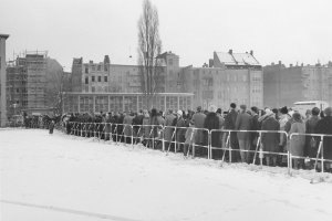 Eine sehr lange Menschenschlange steht entlang eines Absperrgitters im Schnee vor einem Gebäude, das klein im Hintergrund zu erkennen ist.