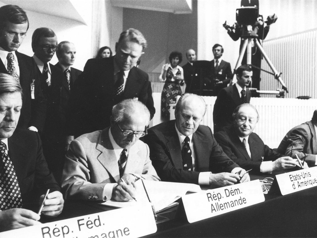 Unterzeichnung der KSZE-Schlussakte in Helsinki am 1. August 1975 (v.l.n.r.: Helmut Schmidt, Erich Honecker, Gerald Ford, Bruno Kreisky). Honecker ist über den Tisch gebeugt und unterschreibt die Akte, die Blicke des US-Präsidenten und des österreichischen Kanzlers sind auf ihn gerichtet. Helmut Schmidt hat den Blick gesenkt. Vor den Regierungschefs stehen Schilder, die mit den französischen Namen ihrer jeweiligen Länder beschriftet sind. Hinter ihnen stehen weitere Menschen und eine Fernsehkamera.