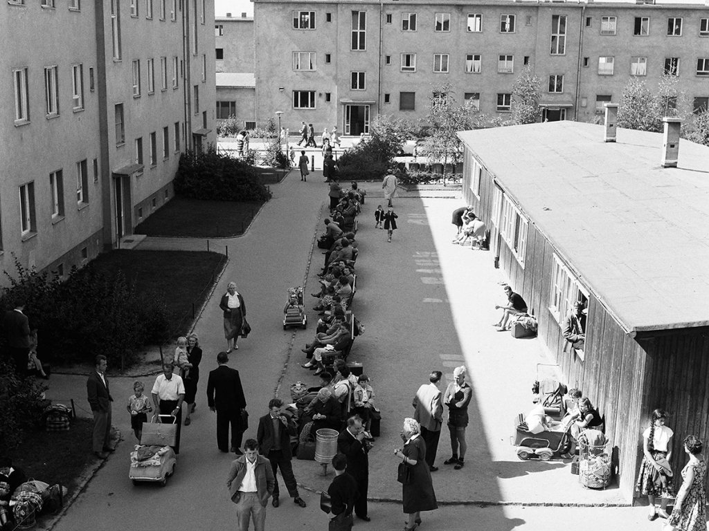 Der Blick geht von oben auf eine Straße, die zwischen zwei mehrstöckigen Gebäuden und einer Baracke liegt. In der Mitte stehen Menschen oder sitzen auf Bänken, einige mit Gepäck und Kinderwägen.