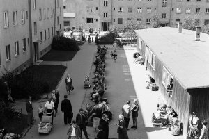 Der Blick geht von oben auf eine Straße, die zwischen zwei mehrstöckigen Gebäuden und einer Baracke liegt. In der Mitte stehen Menschen oder sitzen auf Bänken, einige mit Gepäck und Kinderwägen.