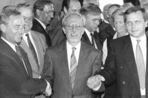 Lothar de Maiziere steht in der Bildmitte mit einem breiten Lächeln. Von links und rechts werden ihm die Hände von Wolfgang Schäuble und Günther Krause gedrückt, die ebenfalls lächeln. Im Hintergrund stehen weitere fröhlich aussehende Menschen.