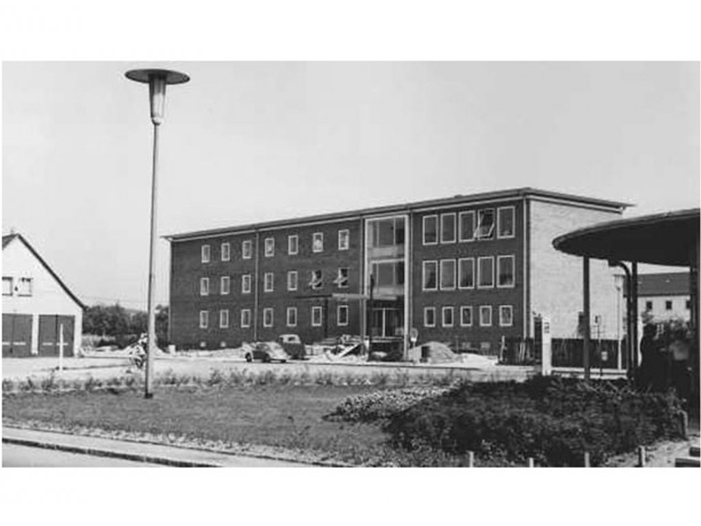 Schwarz-Weiß-Aufnahme. Ein dreigeschossiger Häuserblock steht in der Bildmitte, links ein Giebelhaus, im Vordergrund eine Straßenlaterne und ein Stück Rasen.