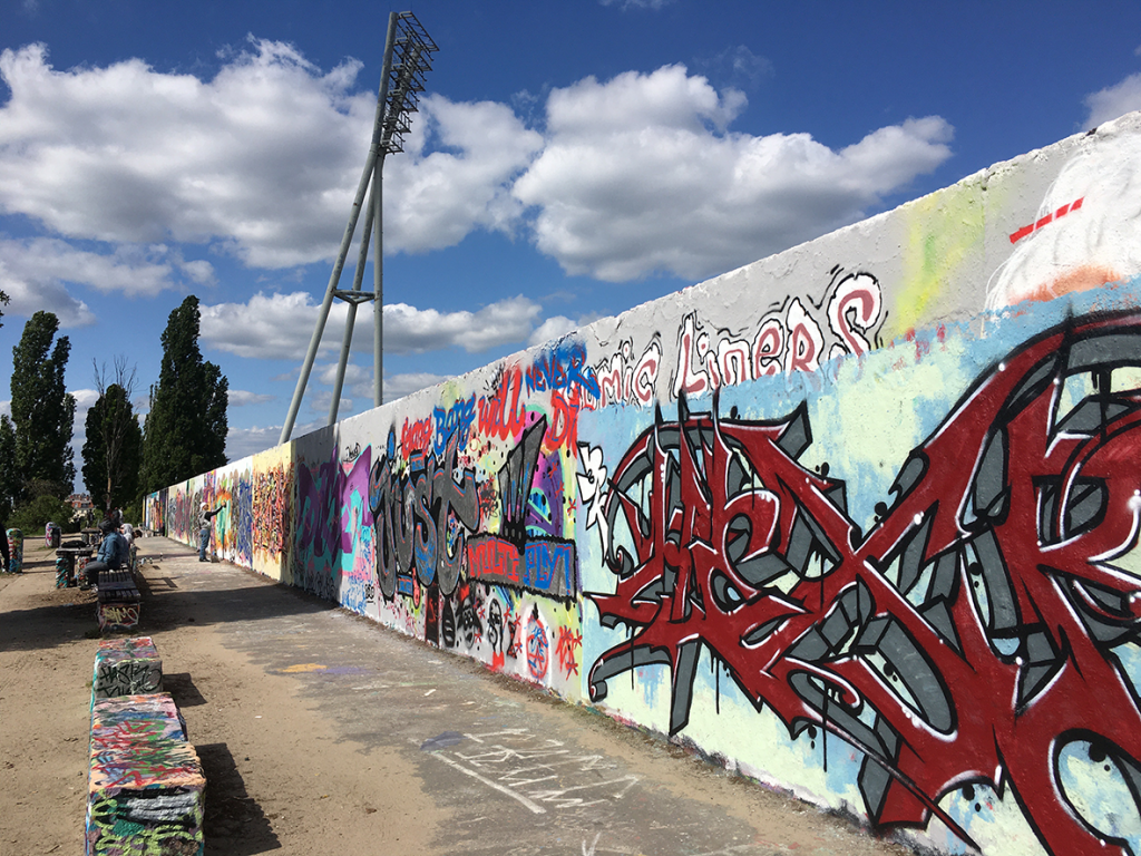Dominant im Bild ist die mit bunten Graffiti bemalte Hinterlandmauer, die ihren Fluchtpunkt mittig am linken Bildrand hat. Vor der Mauer reihen sich buntbemalte steinerne Sitzbänke. Im Bildhintergrund ragt die Flutlichtanlage des Friedrich Ludwig Jahn Sportparks in den Himmel.