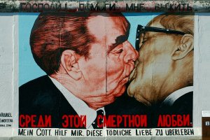 Das Bild zeigt das Gemälde der küssenden Politiker auf der Mauer. Im unteren Bereich steht der Titel des Kunstwerkes auf russisch und auf deutsch.