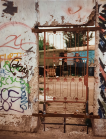 Large section of the Berlin Wall covered in graffiti. In the middle of the wall is a red iron gate allowing a view through the wall and onto two caravans in the background.