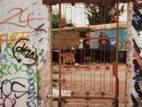 Large section of the Berlin Wall covered in graffiti. In the middle of the wall is a red iron gate allowing a view through the wall and onto two caravans in the background.
