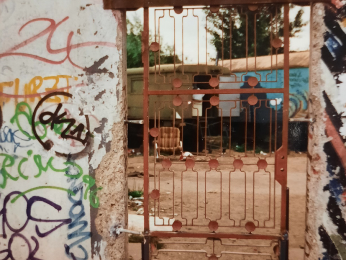 Iron gate in the Berlin Wall with a view of the “East Side Wagenburg,” July 1996 Large section of the Berlin Wall covered in graffiti. In the middle of the wall is a red iron gate allowing a view through the wall and onto two caravans in the background.