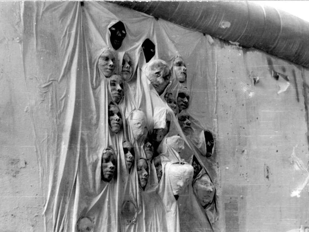 Ein Mauerabschnitt ist mit einem Stofftuch bedeckt. Aus den Falten des Tuches ragen 23 Gipsköpfe.