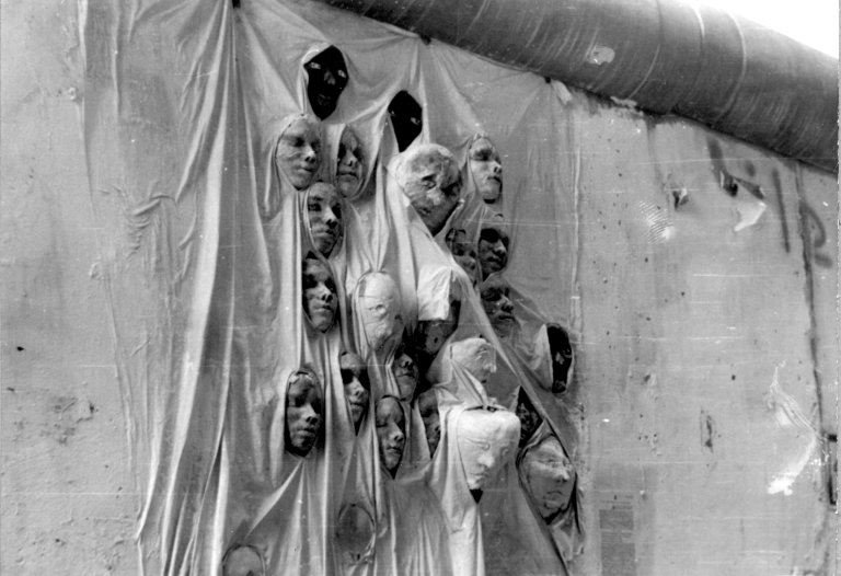 Ein Mauerabschnitt ist mit einem Stofftuch bedeckt. Aus den Falten des Tuches ragen 23 Gipsköpfe.