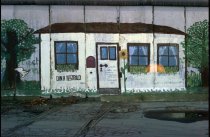 Ein über mehrere Mauersegmente gemaltes Bild zeigt ein weißes Haus zwischen zwei Bäumen. Das Haus hat eine Tür, drei Fenster und einen Briefkasten. Unter dem Fenster links steht: Din A Testbild. Neben der Tür ist eine Sonnenblume gemalt.